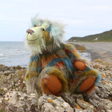Colourful teddy bear sitting on a rocky outcrop looking out to sea, with ocean and beach  in the background