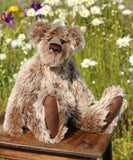 Clitheroe a vintage mohair teddy bear with shaggy silver and brown fur sits on a wooden table in a sunlit flower field. The bear has dark brown paws and a dark brown snout, with a blurred background of green grass and white, yellow, and purple wildflowers.