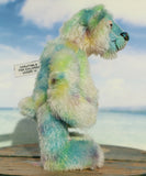 Profile of Colin Cockles a colourful hand dyed mohair teddy bear standing on a wooden surface with a warning label against a beach background