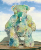 Back view of Colin Cockles a colourful hand dyed mohair teddy bear sitting on a wooden surface with a blurred beach background