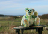 Colin Cockles a colourful hand dyed mohair teddy bear sitting on a wooden bench in a field with a blurred background