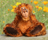 Denzel, a happy brown tipped ginger mohair artist bear by Barbara A Bears, photographed sitting on a wooden surface in front of a blurred green field with yellow and orange poppies.