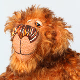 A close-up of Denzel's face, a happy brown tipped ginger mohair artist bear by Barbara A Bears, photographed sitting against a plain light grey background.