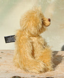 Euan sitting in profile a mohair collector's teddy bear with a soft texture and with a safety warning tag against a light background  on a wooden surface