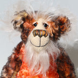 A close up of Flambé by Barbara Ann Bears, a handmade, distressed teddy bear with a long fluffy mohair face, with hand painted eyes, a multi coloured nose and a beaming smile. Made from shaggy brown and orange fur, a cream-coloured stitched face