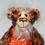 A close up of Flambé by Barbara Ann Bears, a handmade, distressed teddy bear with a long fluffy mohair face, with hand painted eyes, a multi coloured nose and a beaming smile. Made from shaggy brown and orange fur, a cream-coloured stitched face