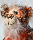 A close up of Flambé by Barbara Ann Bears, a handmade, distressed teddy bear with a long fluffy mohair face, with hand painted eyes, a multi coloured nose and a beaming smile. Made from shaggy brown and orange fur, a cream-coloured stitched face
