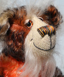 A close up of Flambé by Barbara Ann Bears, a handmade, distressed teddy bear with a long fluffy mohair face, with hand painted eyes, a multi coloured nose and a beaming smile. Made from shaggy brown and orange fur, a cream-coloured stitched face