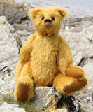 Fortescue, a golden teddy bear by Barbara Ann Bears with shaggy fur sits on a rocky, grassy shoreline near Lochranza on the Isle of Arran, Scotland.