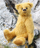 Fortescue, a golden teddy bear by Barbara Ann Bears with shaggy fur sits on a rocky, grassy shoreline near Lochranza on the Isle of Arran, Scotland.