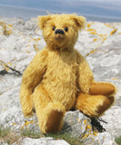 Fortescue, a golden teddy bear by Barbara Ann Bears with shaggy fur sits on a rocky, grassy shoreline near Lochranza on the Isle of Arran, Scotland.
