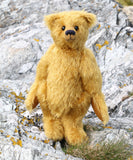 Fortescue, a golden teddy bear by Barbara Ann Bears with shaggy fur stands on a rocky, grassy shoreline near Lochranza on the Isle of Arran, Scotland.