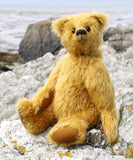Fortescue, a golden teddy bear by Barbara Ann Bears with shaggy fur sits on a rocky,  shoreline near Lochranza on the Isle of Arran, Scotland.