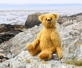 Fortescue, a golden teddy bear by Barbara Ann Bears with shaggy fur sits on a rocky, grassy shoreline near Lochranza on the Isle of Arran, Scotland, with water and the distant hills of The Mull of Kintyre in the background.