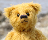 A close-up of Fortescue, a fluffy golden-yellow teddy bear, by Barbara An Bears, with black eyes and a dark nose, photographed against a softly blurred outdoor background.