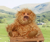 Plush teddy bear on a wooden stool with a scenic background of green fields and mountains.
