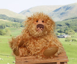 Plush teddy bear on a wooden stool with a scenic background of green fields and mountains.