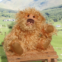 Plush teddy bear on a wooden stool with a scenic background of mountains and fields.