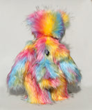 Cock A Doodle Doo viewed from the back, a colourful, furry teddy bear with a shaggy, multi-hued coat in pink, yellow, blue, orange, and green, standing against a plain light grey background.