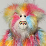 A close-up of Cock A Doodle Doo's face, a fluffy teddy bear with a rainbow fur pattern, featuring a white mohair face with hand painted glass eyes, a multicoloured nose and a sweet smile set against a plain light grey background.  