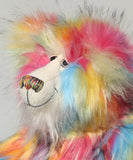 A close-up of Cock A Doodle Doo's face, a fluffy teddy bear with a rainbow fur pattern, featuring a white mohair face with hand painted glass eyes, a multicoloured nose and a sweet smile set against a plain light grey background.  