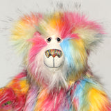 A close-up of Cock A Doodle Doo's face, a fluffy teddy bear with a rainbow fur pattern, featuring a white mohair face with hand painted glass eyes, a multicoloured nose and a sweet smile set against a plain light grey background.  