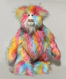 Cock A Doodle Doo, a colourful, furry teddy bear with a shaggy, multi-hued coat in pink, yellow, blue, orange, and green, standing against a plain light grey background.